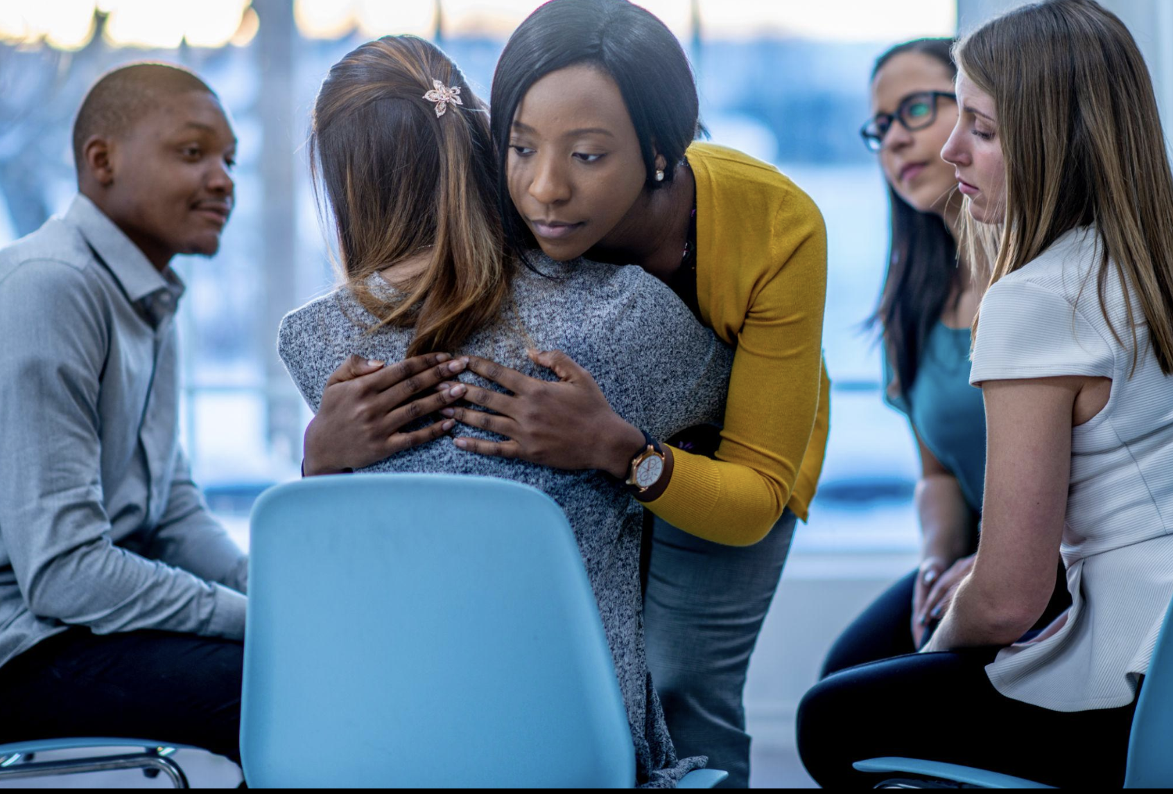 A small group setting where one person gently hugs and comforts another while others sit nearby offering support.